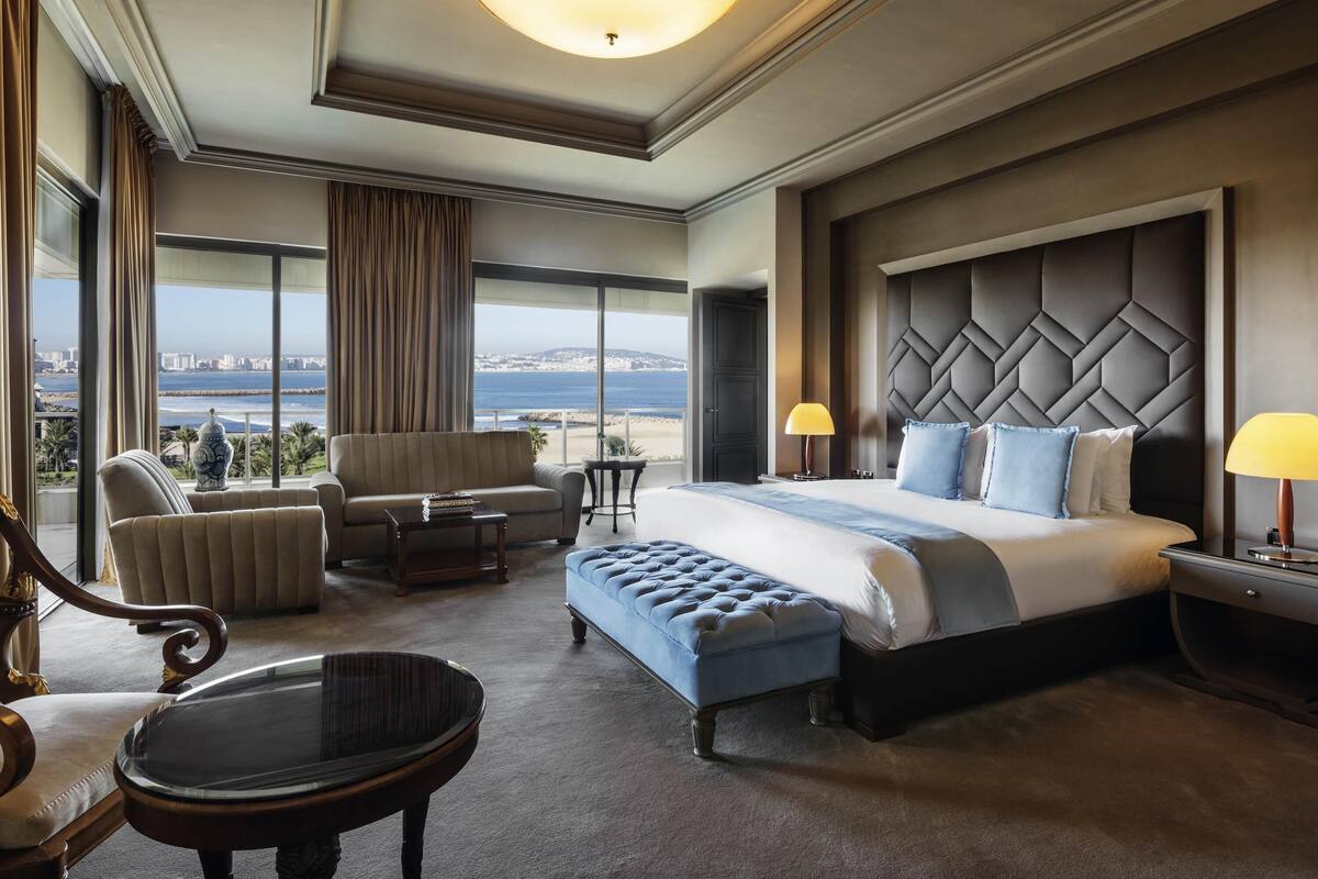 A hotel room with a large bed, a sofa, a chair, a small table, and a view of the sea.
