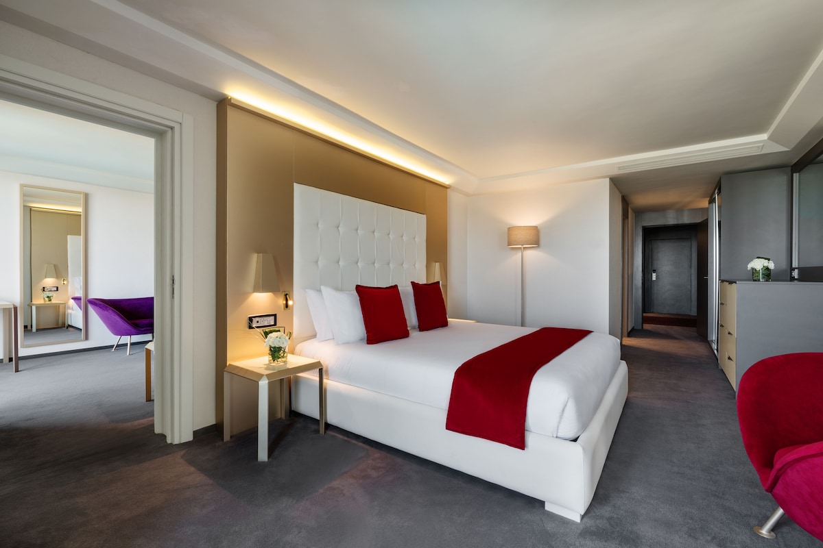 A modern hotel room with a large bed, a bedside table, a lamp, and a mirror.