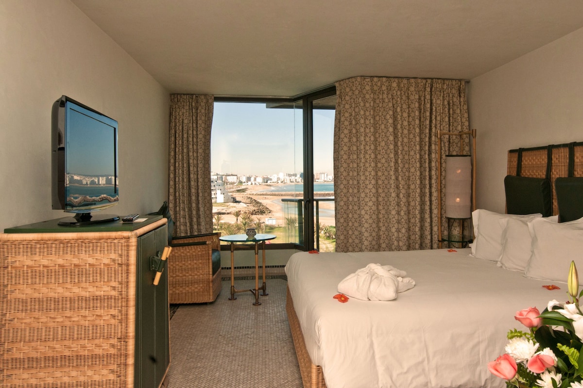 A hotel room with a bed, a TV, a small table, and a view of the city.
