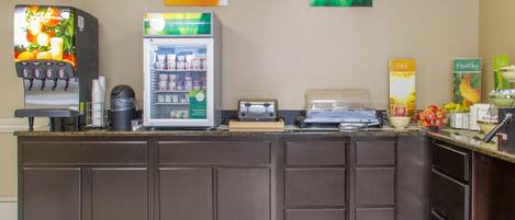 A breakfast bar with a coffee machine, a refrigerator stocked with beverages, and a fruit bowl.