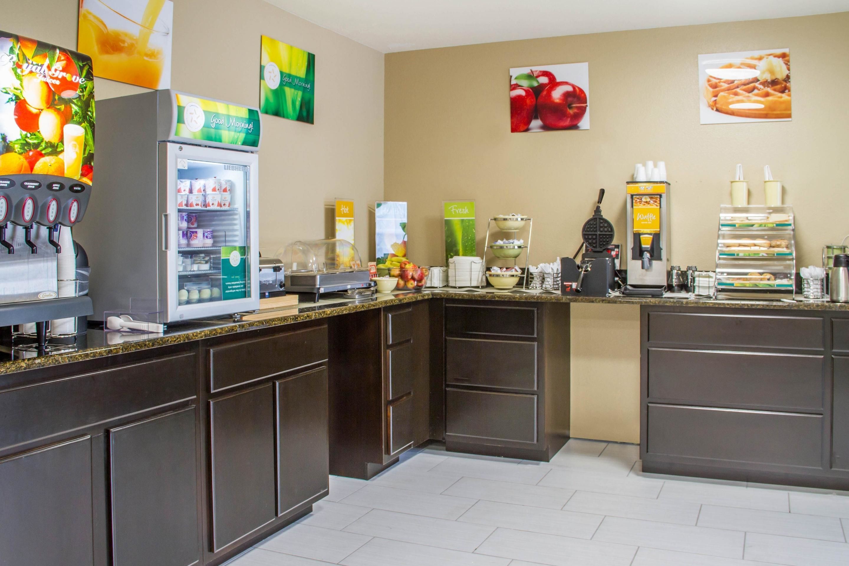 A modern hotel kitchen with a self-service breakfast bar, a refrigerator stocked with beverages, and a coffee machine.