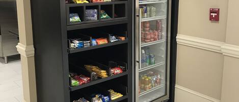 A vending machine stocked with snacks and beverages, located in a hallway with a tiled floor and a fire alarm on the wall.