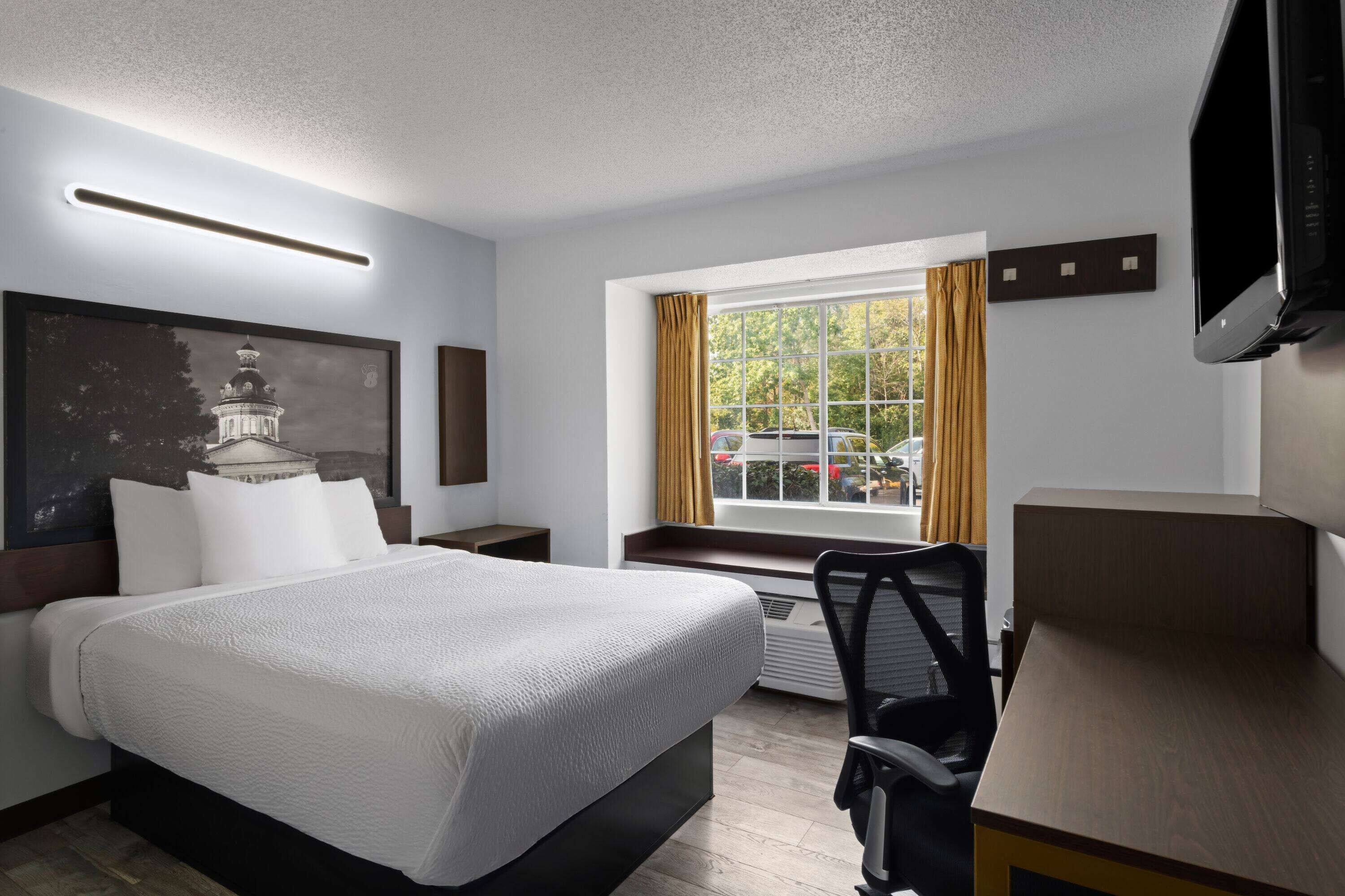 Quarto de hotel com uma cama grande, uma escrivaninha, uma cadeira, uma televisão, uma janela com cortinas e um quadro na parede.