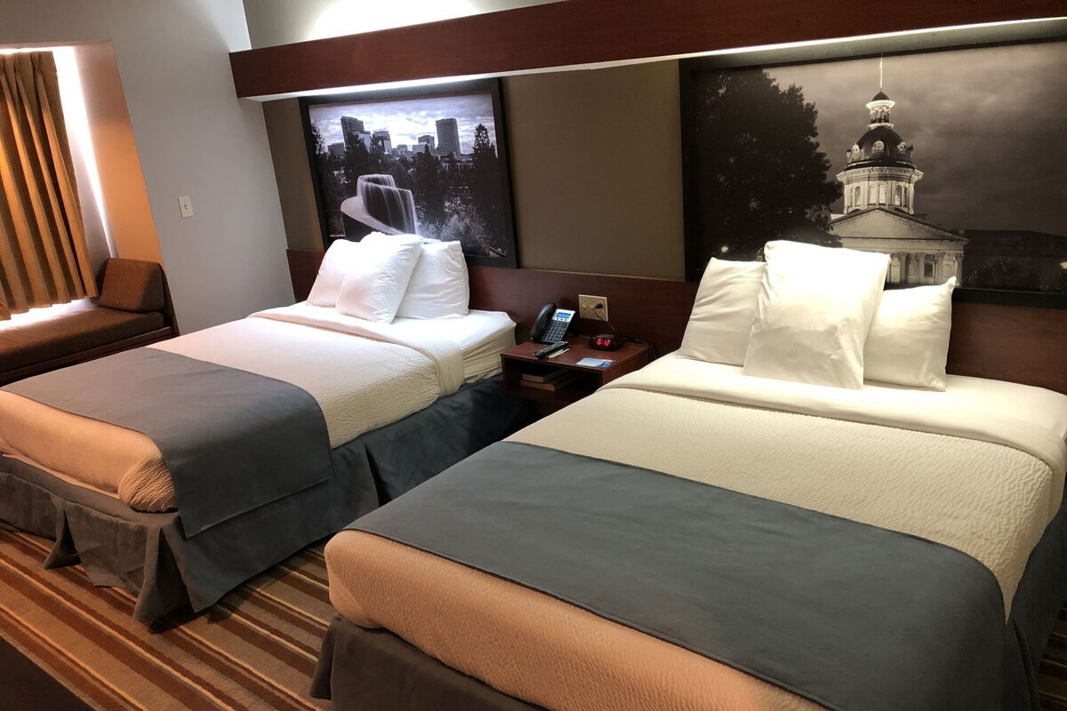 A hotel room with two beds, a nightstand with a phone, and a cityscape painting on the wall.