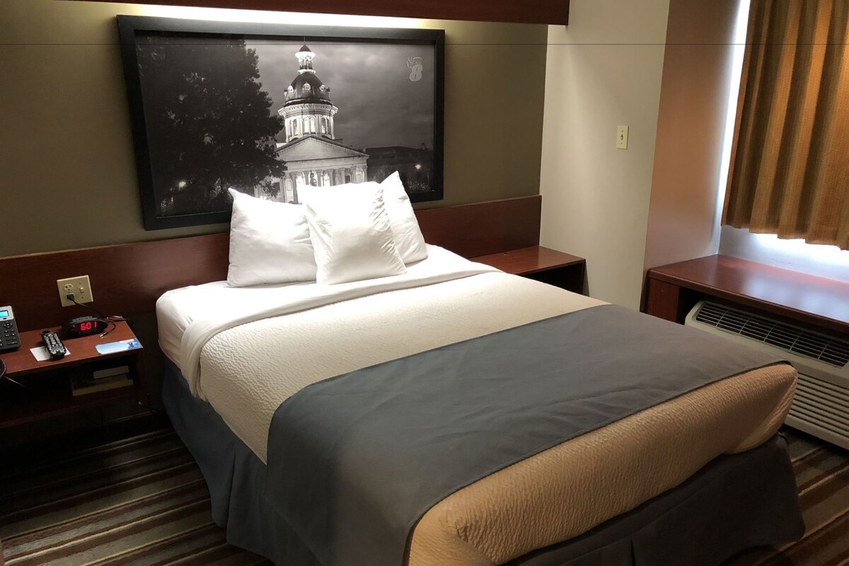 A hotel room with a bed, a nightstand with a phone, a wall-mounted TV, and a window with curtains.