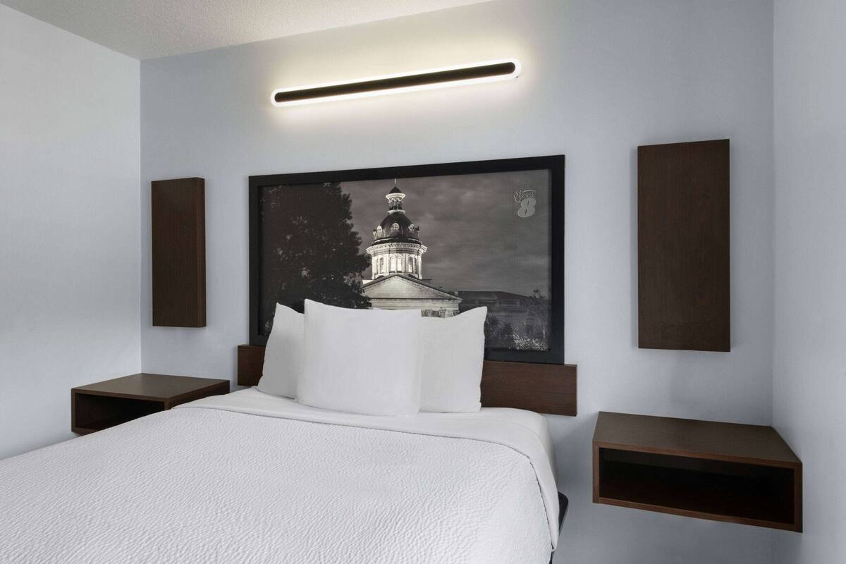 Una habitación de hotel moderna con una cama grande, un cabecero con un cuadro enmarcado, dos mesitas de noche y un aplique de pared.