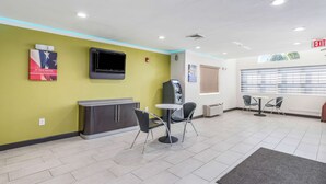 Una hall con una reception, un televisore a parete, un chiosco self-service e un'area salotto con sedie e un tavolo.