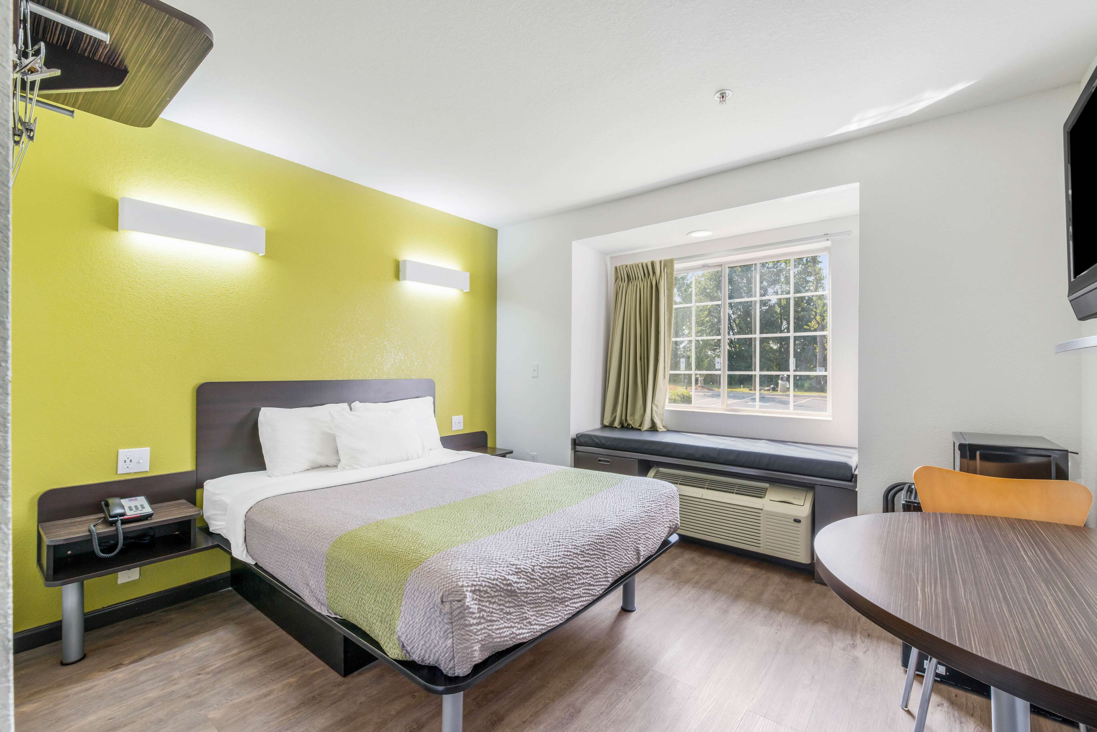 A hotel room with a bed, bedside table, lamp, window with curtains, and a dining table.