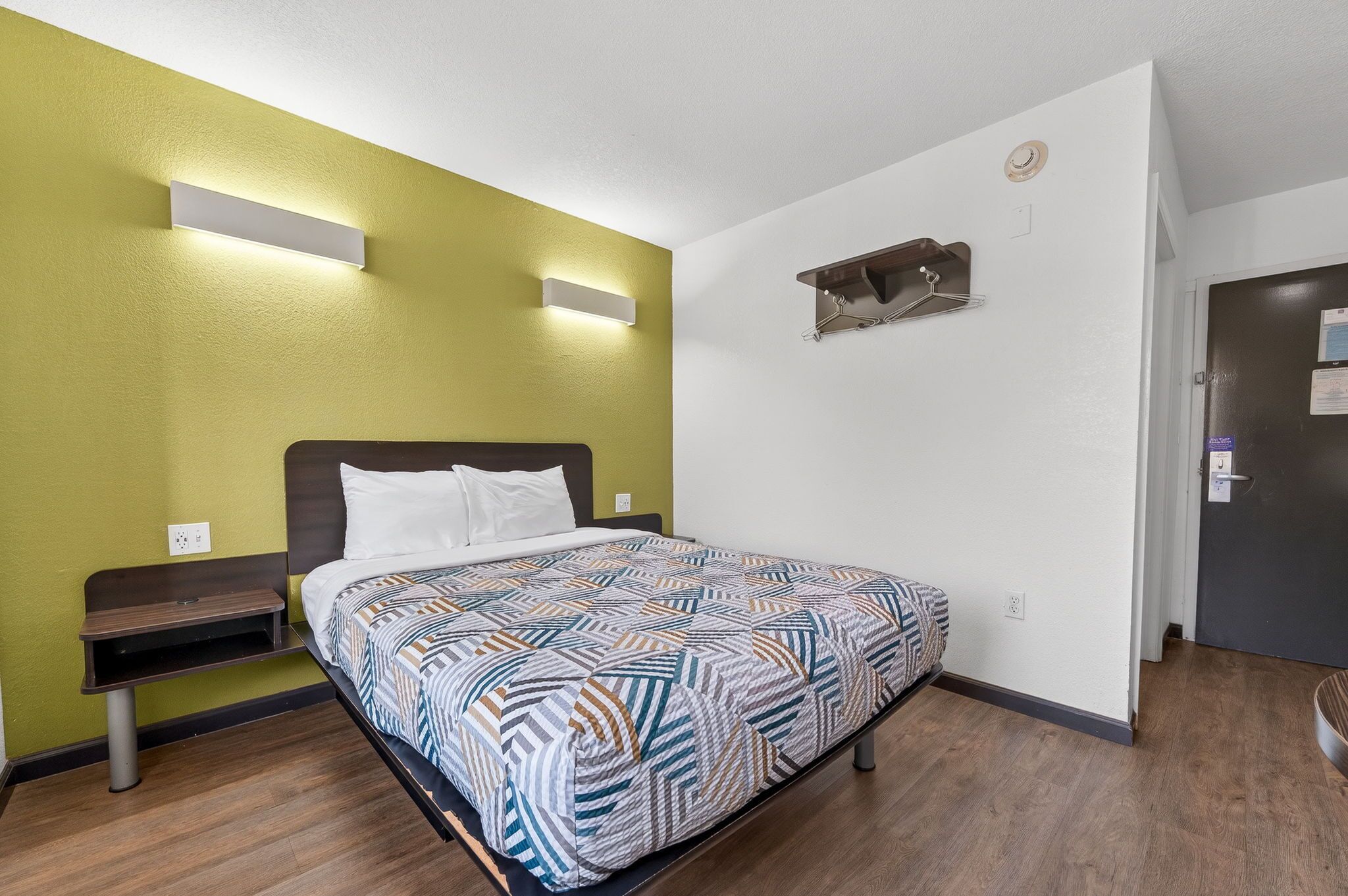 A neatly made bed with a patterned bedspread, a bedside table with a lamp, and a wall-mounted shelf.