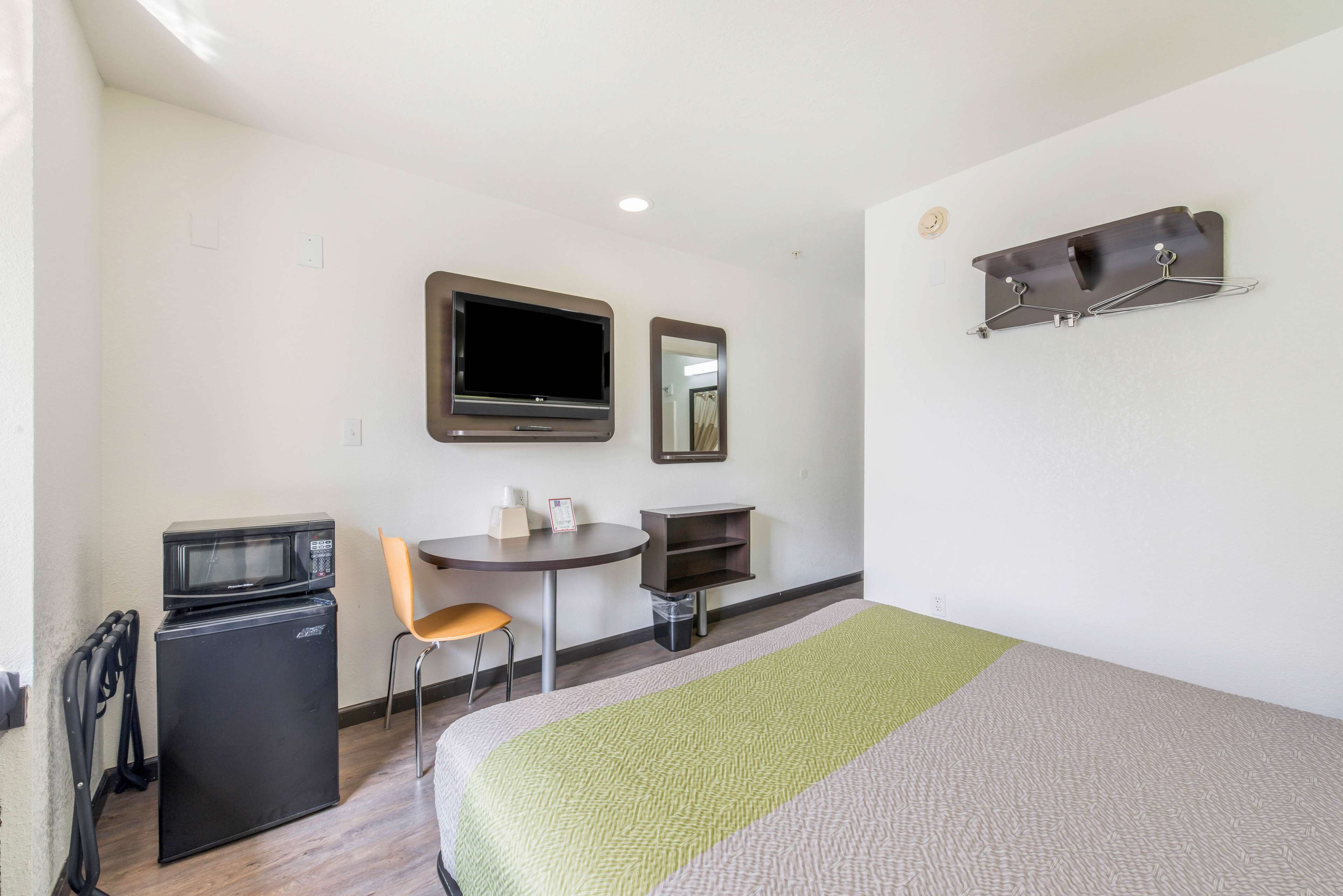 A compact hotel room with a bed, a small desk with a chair, a microwave, a TV, and a mirror.