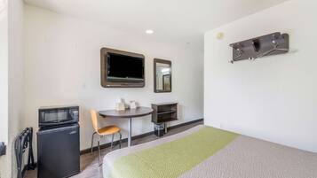 A compact hotel room with a bed, a small desk with a chair, a microwave, a TV, and a mirror.