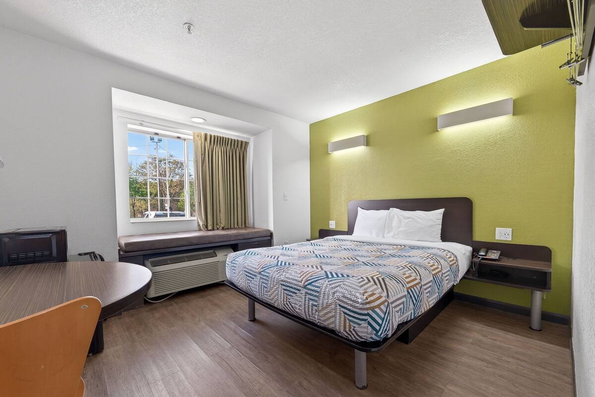 Hotellihuone, jossa on sänky, työpöytä, tuoli, ikkuna verhoilla ja näkymä puita kohti.