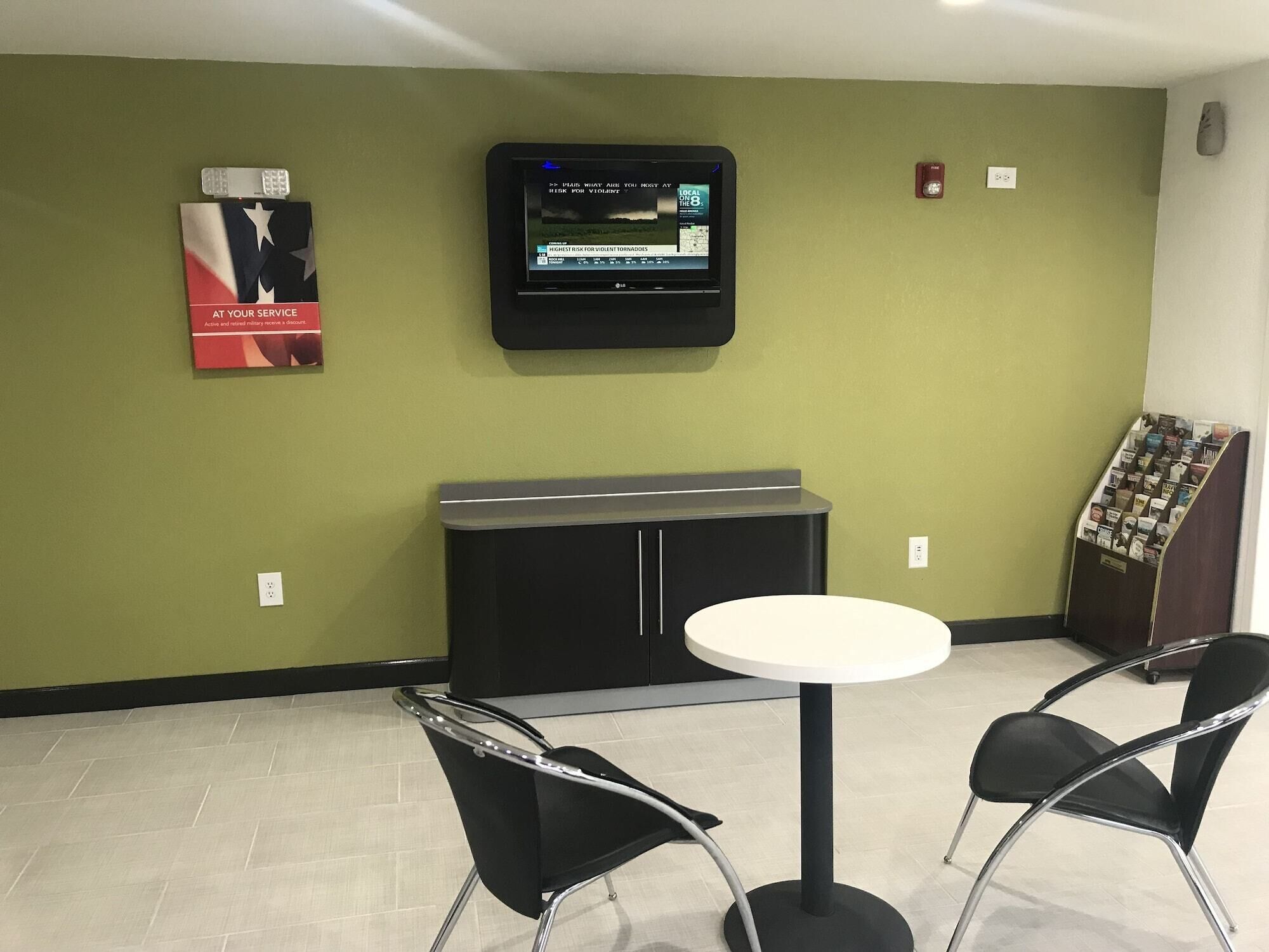 A waiting area with a television mounted on the wall displaying a news channel, a small table with two chairs, and a magazine rack.