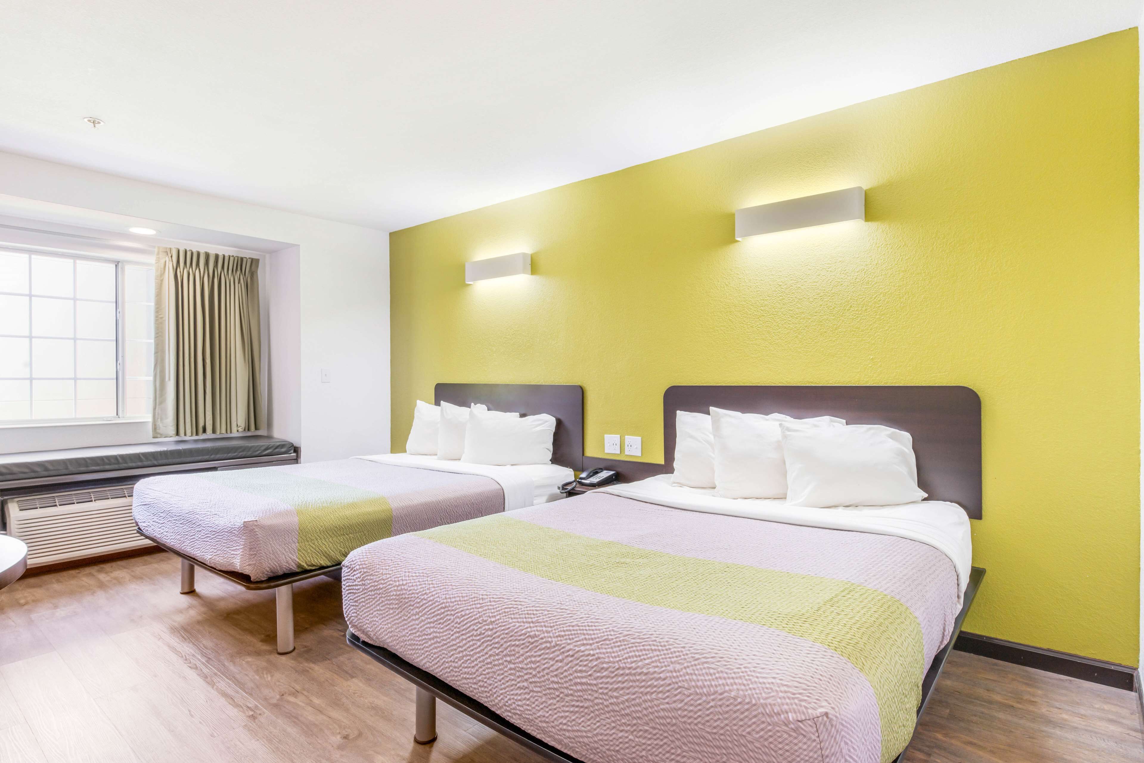 A hotel room with two beds, a window with curtains, a heater, and a wall-mounted phone.