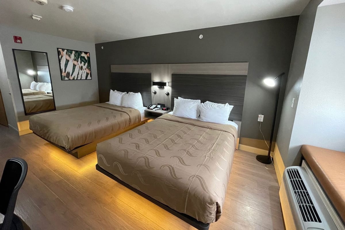 Une chambre d’hôtel avec deux lits, un bureau, une chaise, un miroir et un luminaire fixé au mur.