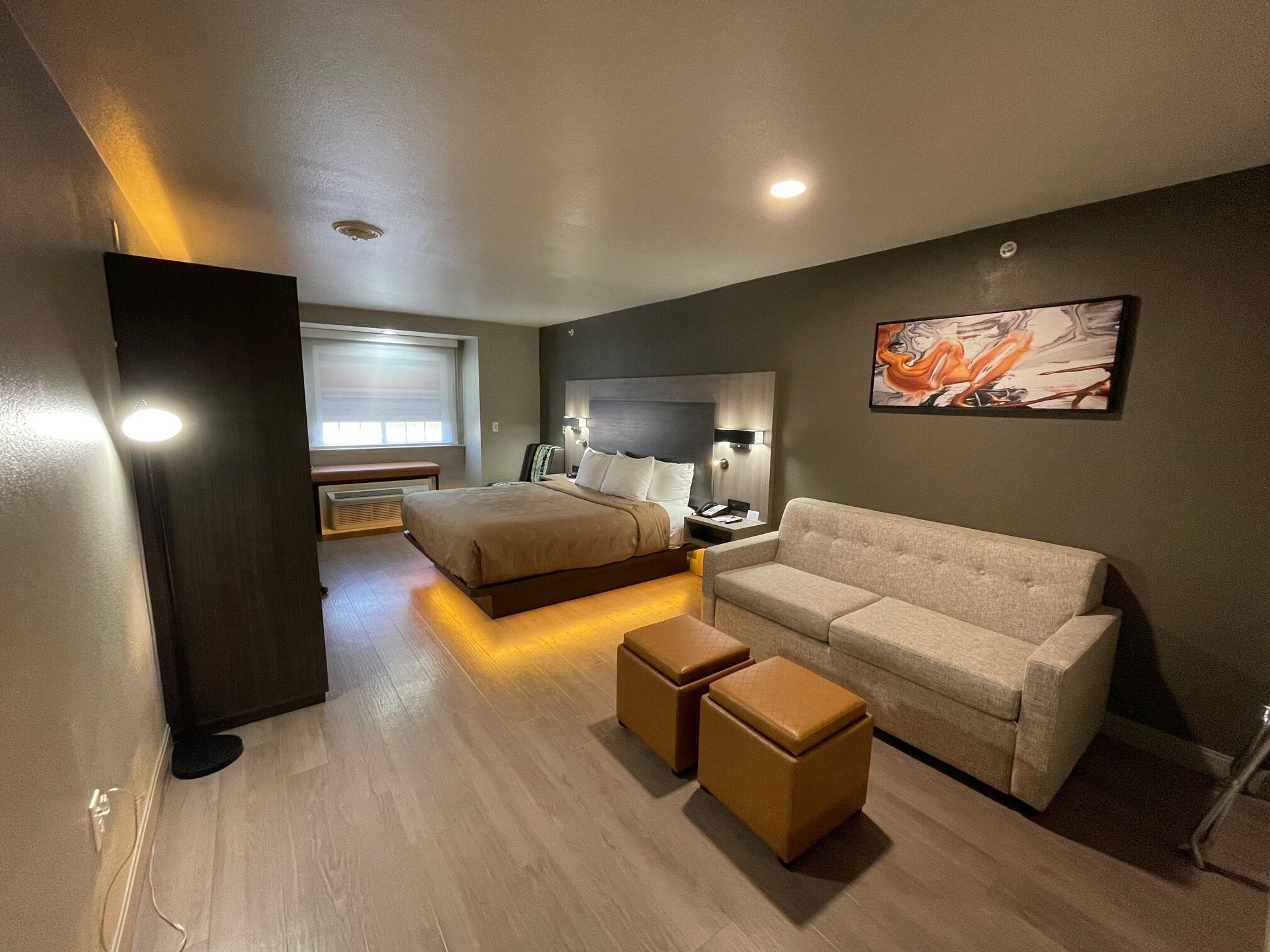 A modern hotel room with a large bed, a sofa, two ottomans, a desk, and a painting on the wall.