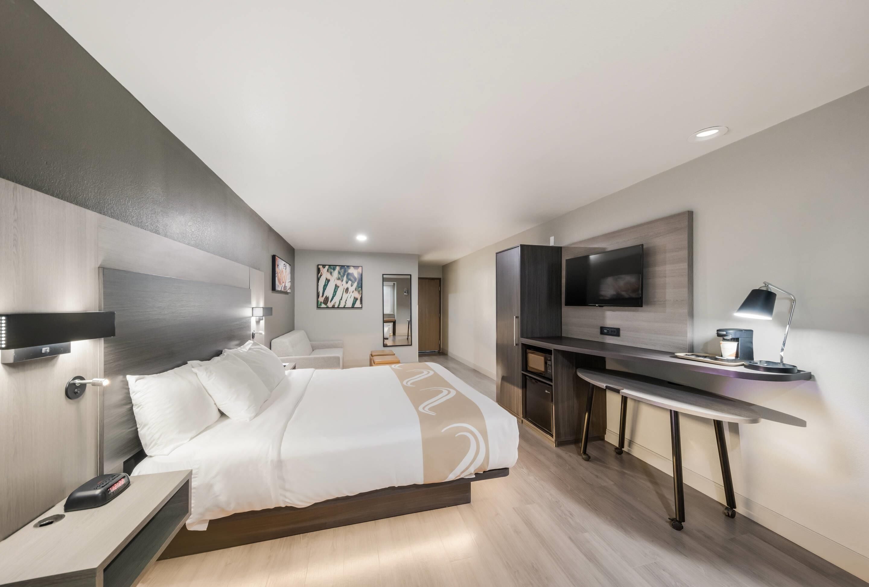 A modern hotel room with a bed, bedside table, desk, and TV.
