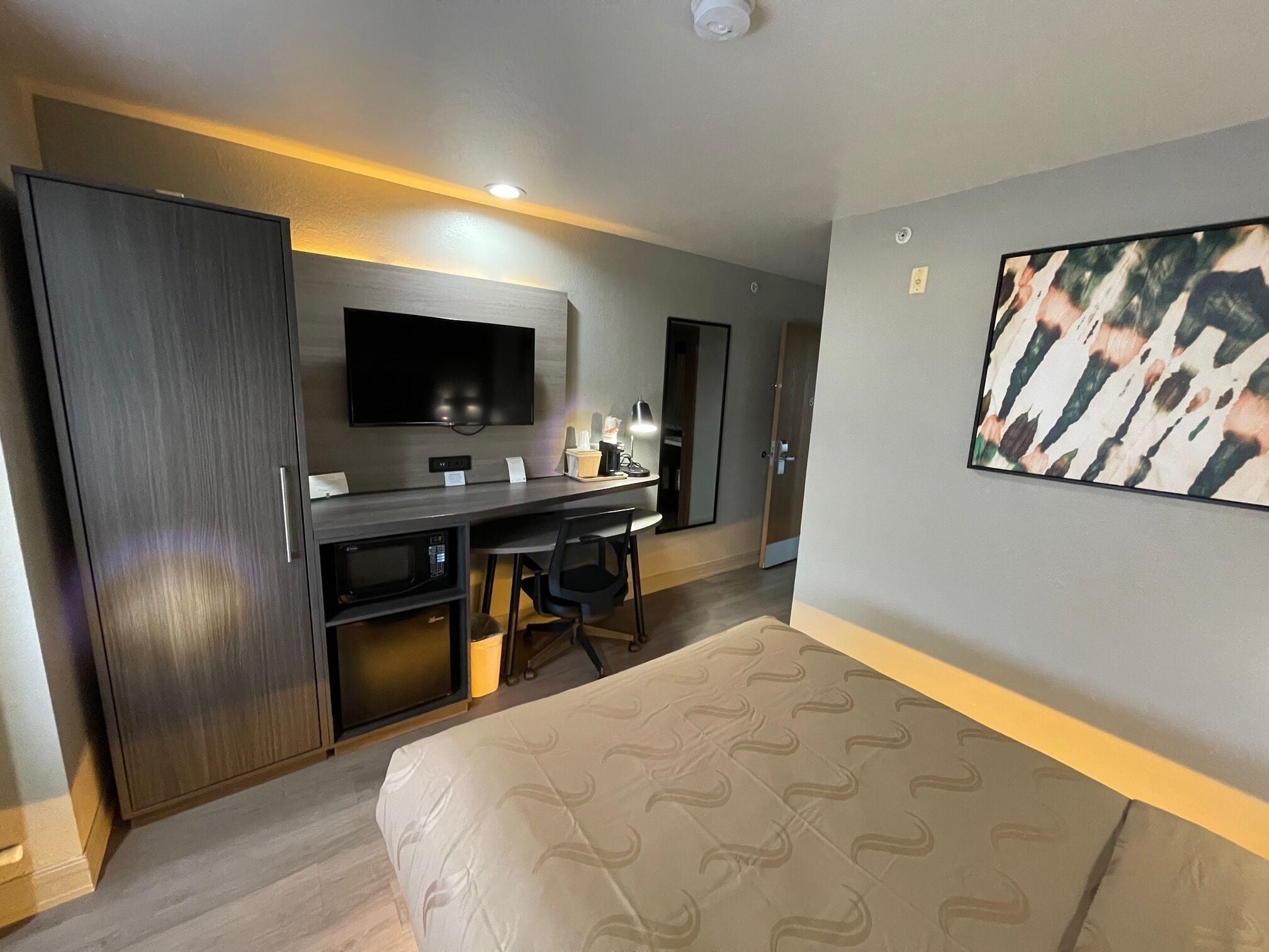 A hotel room with a bed, a desk with a chair, a television, and a painting on the wall.