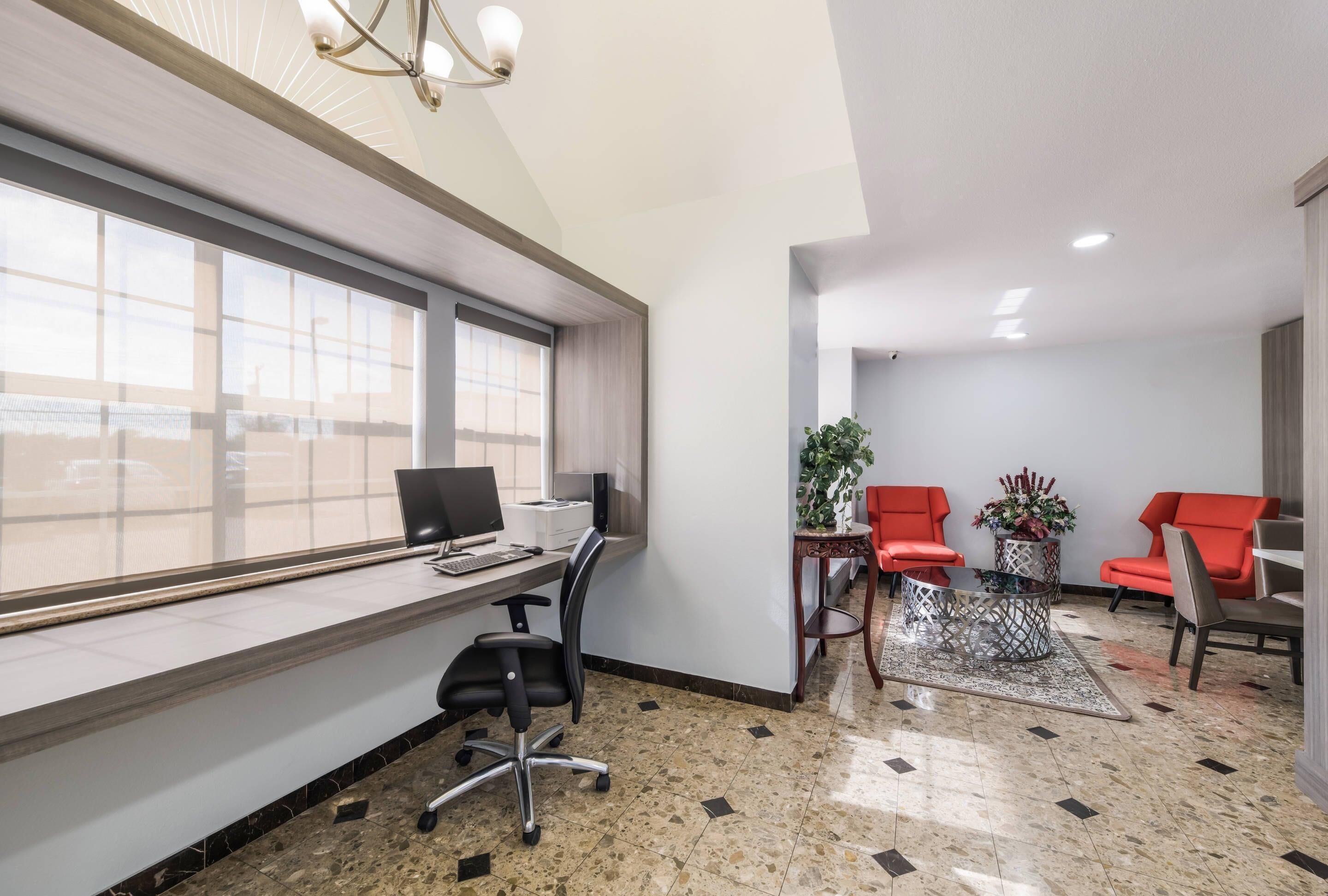 Een moderne kantoorruimte met een bureau, computer en bureaustoel. Er staan twee rode stoelen en een kleine tafel met een plant. De ruimte heeft grote ramen en een betegelde vloer.