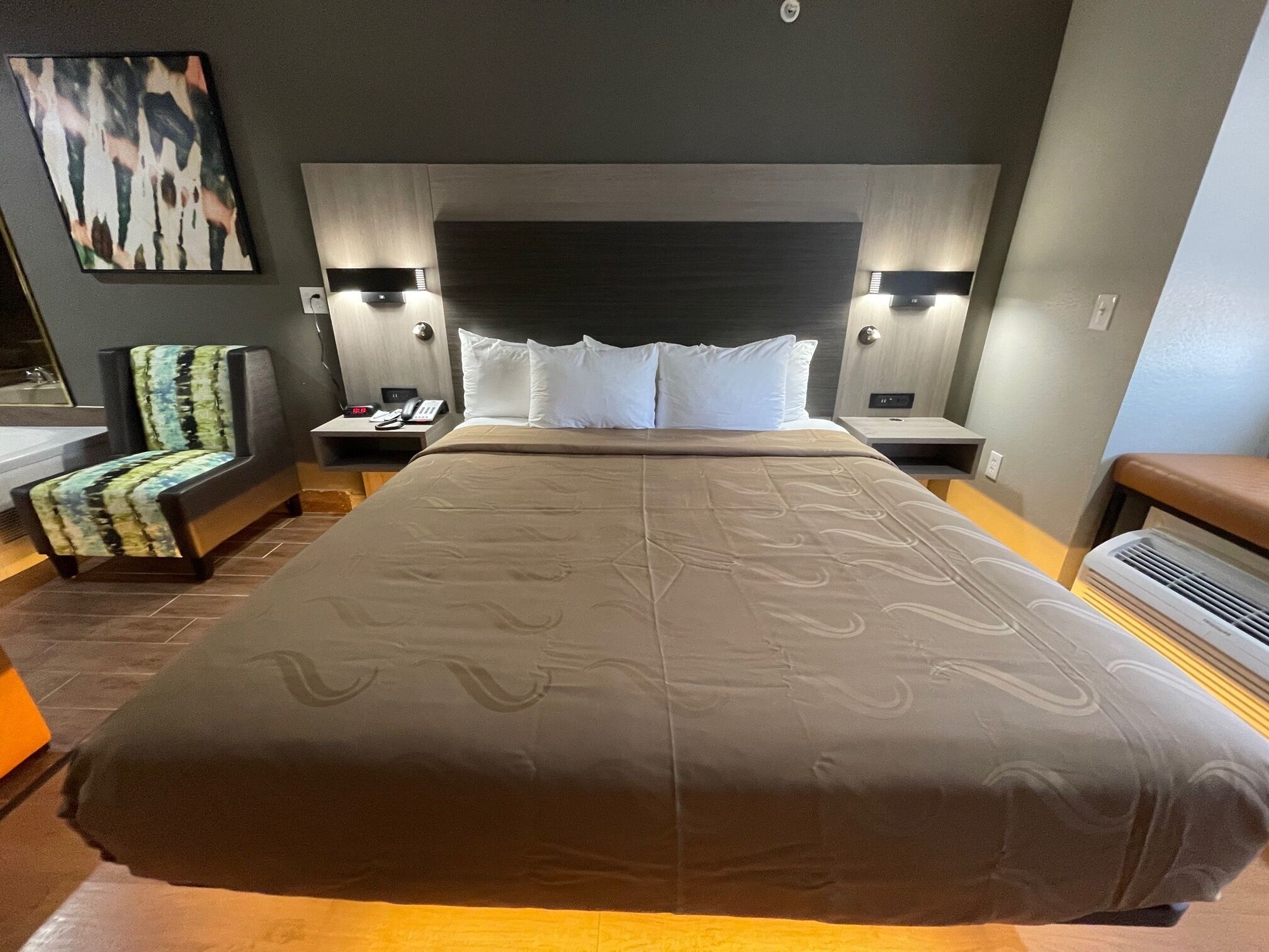 A hotel room with a large bed, a chair, a nightstand, and a wall-mounted light fixture.