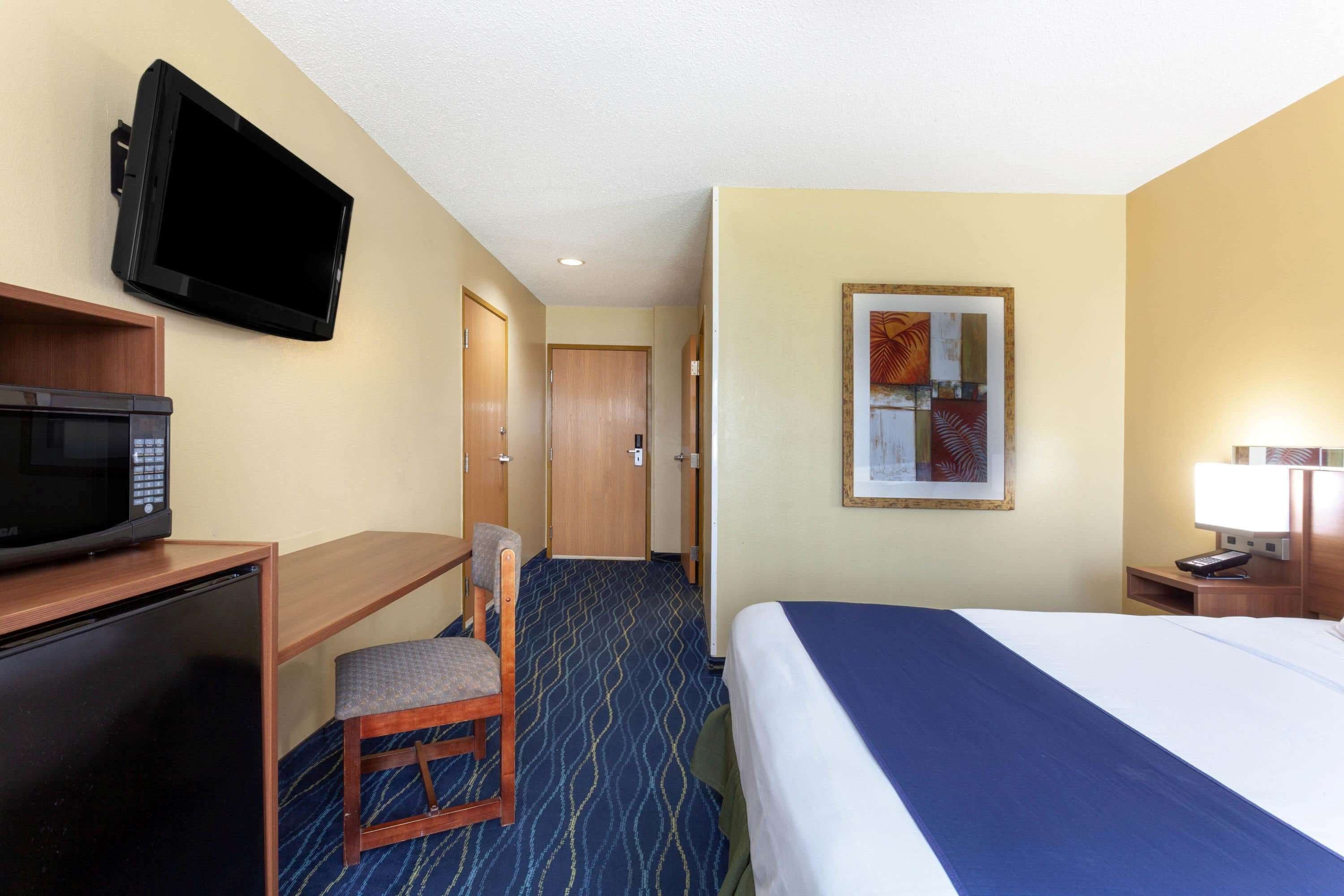 A hotel room with a bed, a desk with a microwave, a television mounted on the wall, and a framed picture on the wall.