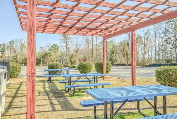 Un área de picnic techada con mesas y bancos de metal, rodeada de árboles y un estacionamiento.