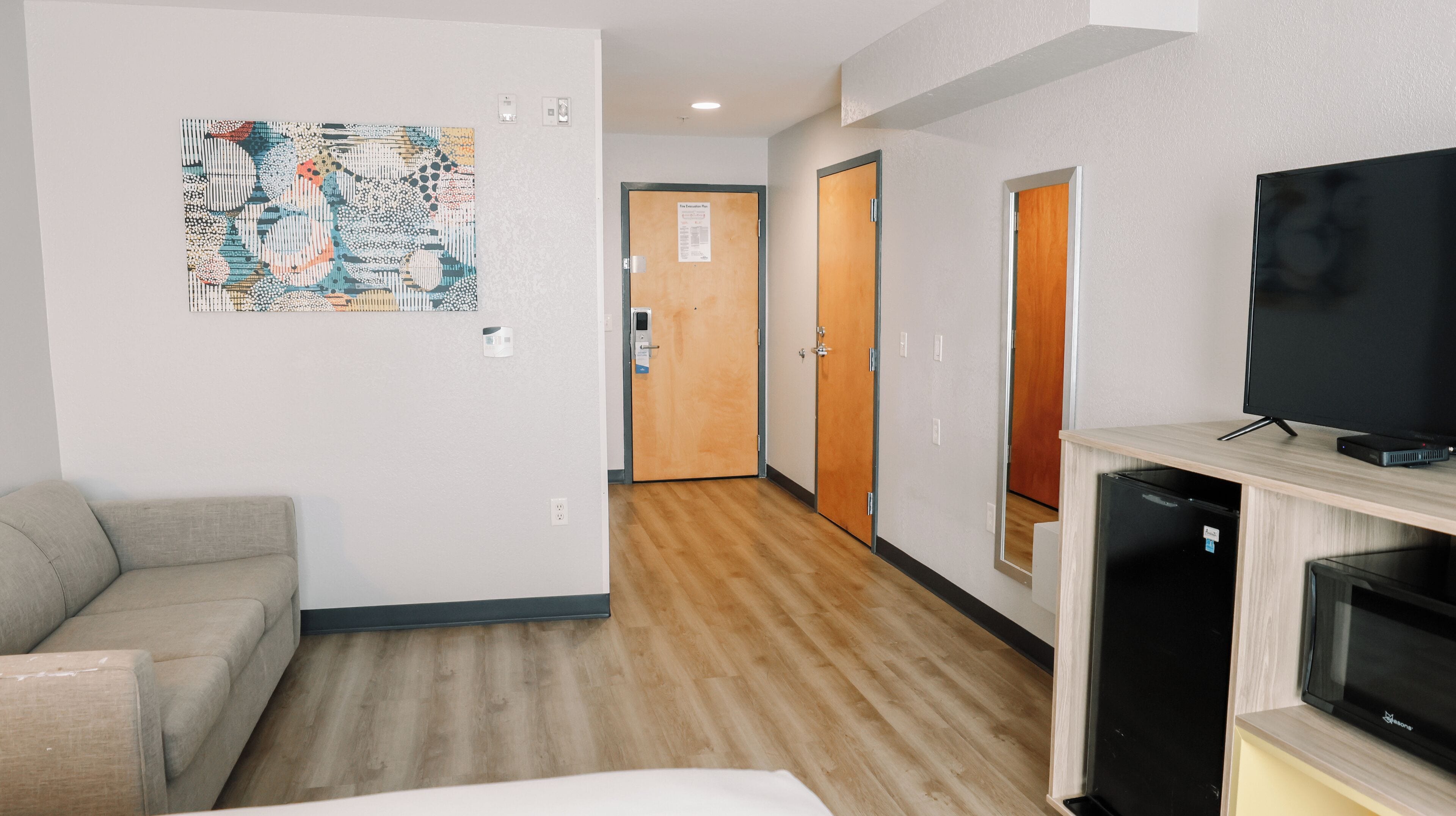 Una habitación de hotel con sofá, televisor, refrigerador y un pasillo con puertas.