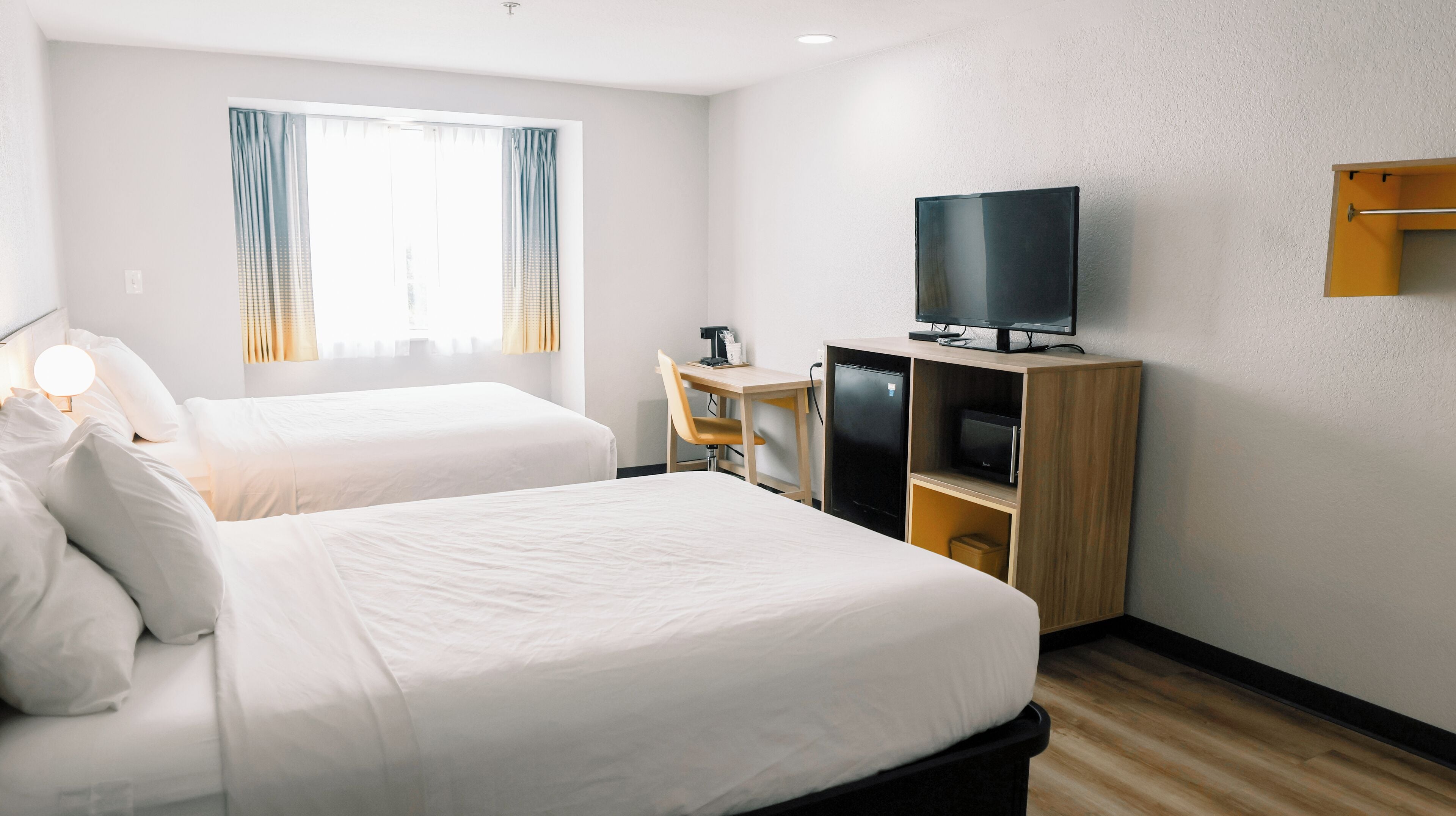 A hotel room with two beds, a desk with a chair, a television on a wooden stand, and a window with curtains.