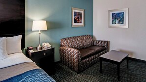 A hotel room with a bed, a nightstand with a lamp, a patterned sofa, a small table, and two framed pictures on the wall.