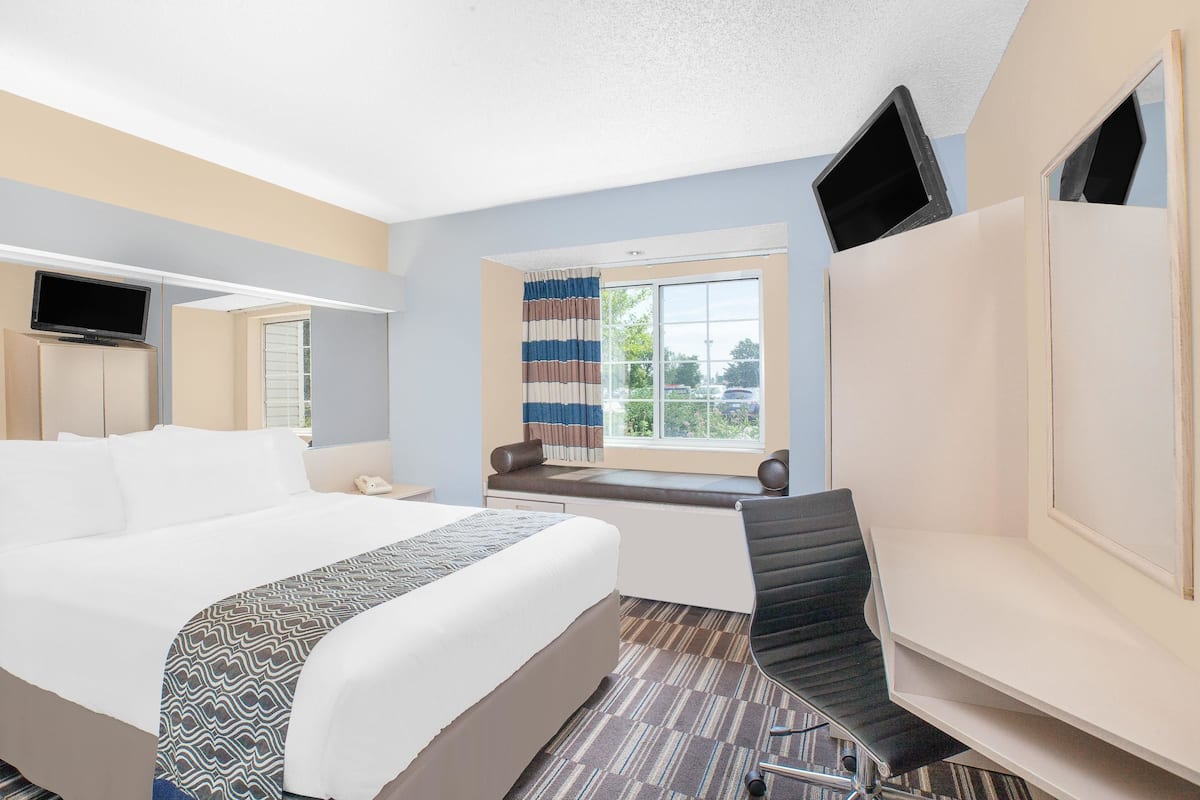 A hotel room with a large bed, a desk, a chair, a TV, a window with curtains, and a view of the outside.