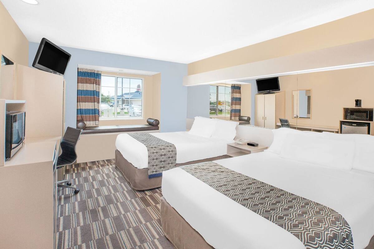 A hotel room with two beds, a TV, a desk, a chair, a microwave, and a window with curtains.