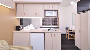 A compact kitchenette with a microwave, a coffee maker, and a small refrigerator. There is a chair and a desk with a chair in the background.