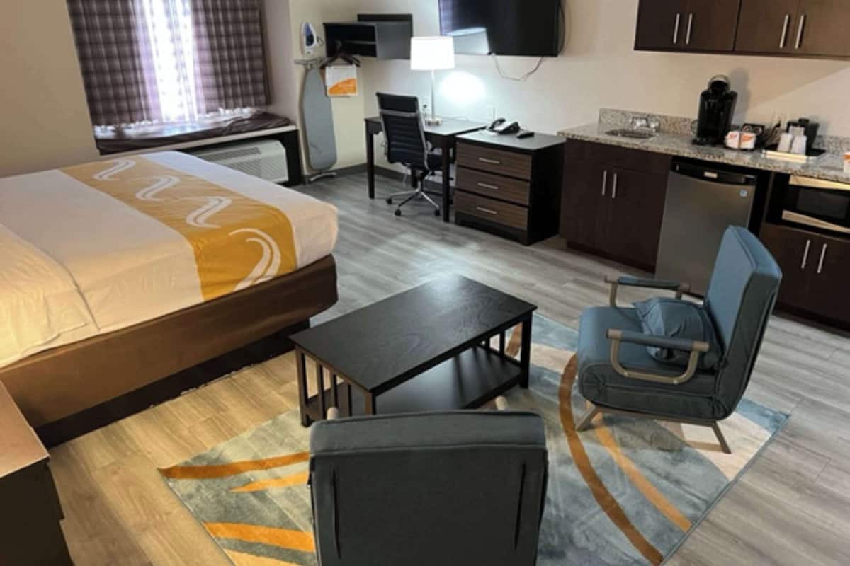 A hotel room with a bed, a desk, a chair, a small table, a TV, a coffee machine, and a kitchen area.