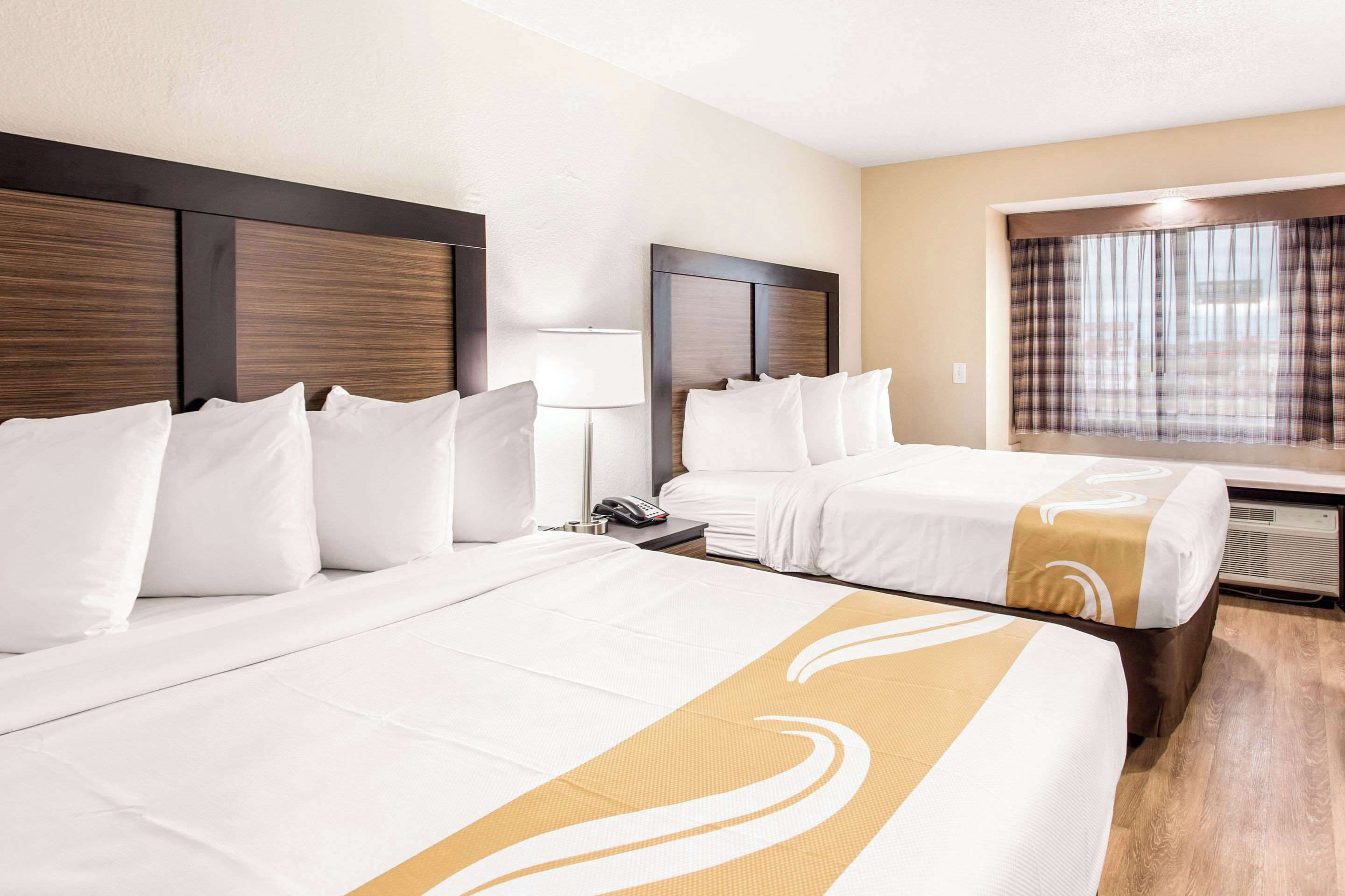 A hotel room with two beds, a bedside table with a lamp, a phone, and a window with curtains.