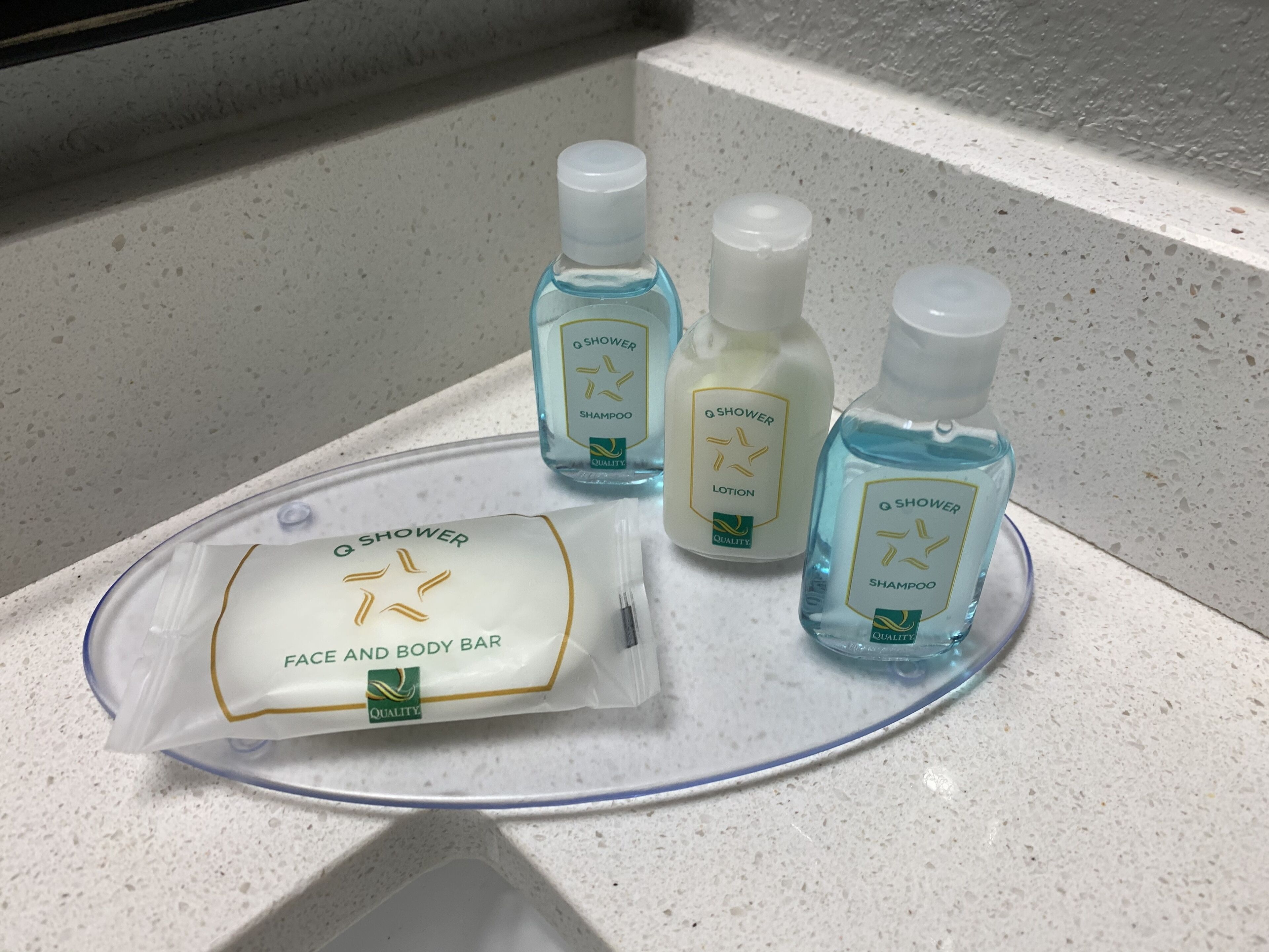 Un lavabo de salle de bain avec un plan-vasque blanc et un porte-savon transparent. Sur le lavabo, on trouve trois flacons de produits de la marque Q Shower : deux flacons de shampoing et un flacon de lotion.