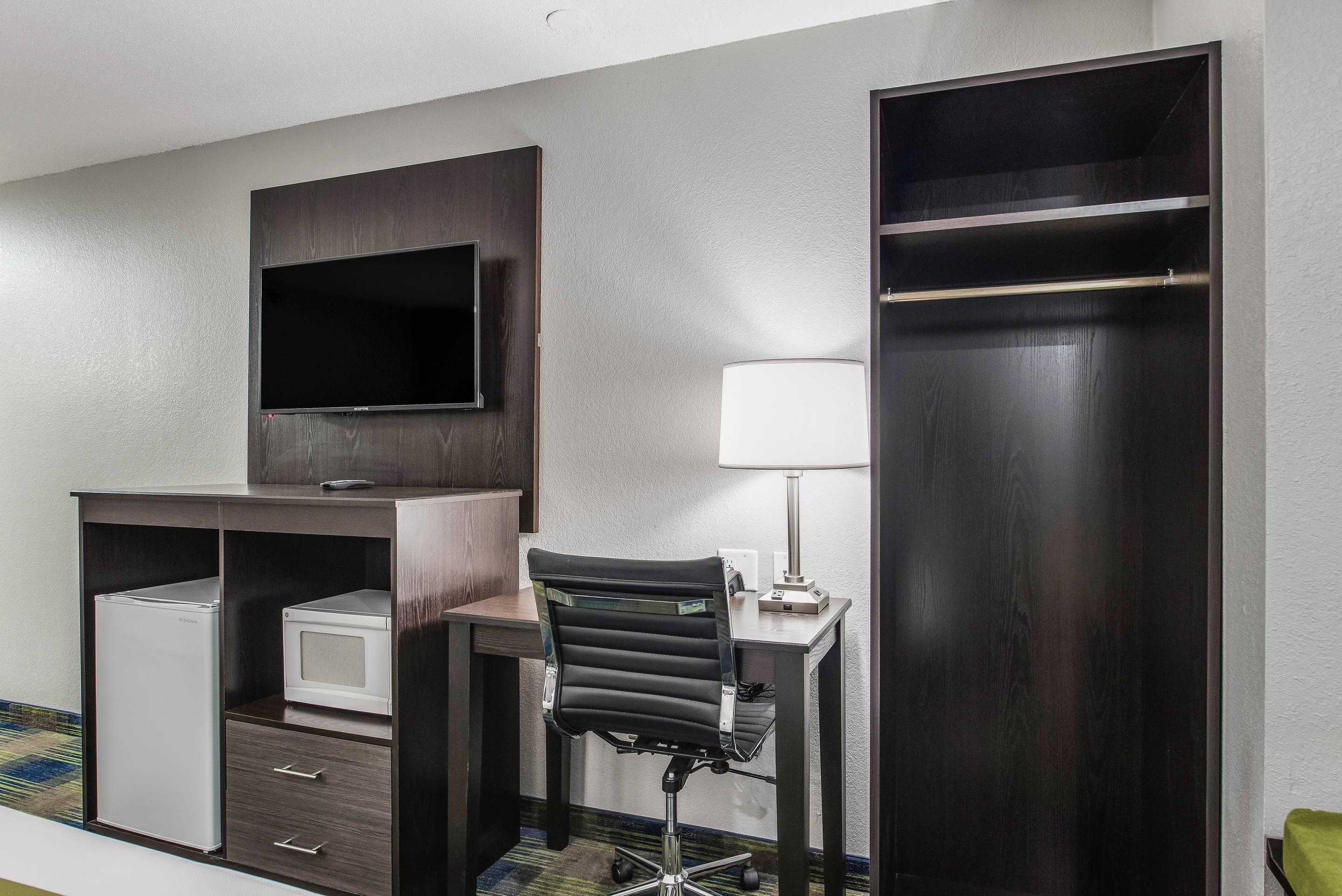 A hotel room with a desk, a chair, a television, a lamp, a wardrobe, and a mini-fridge.