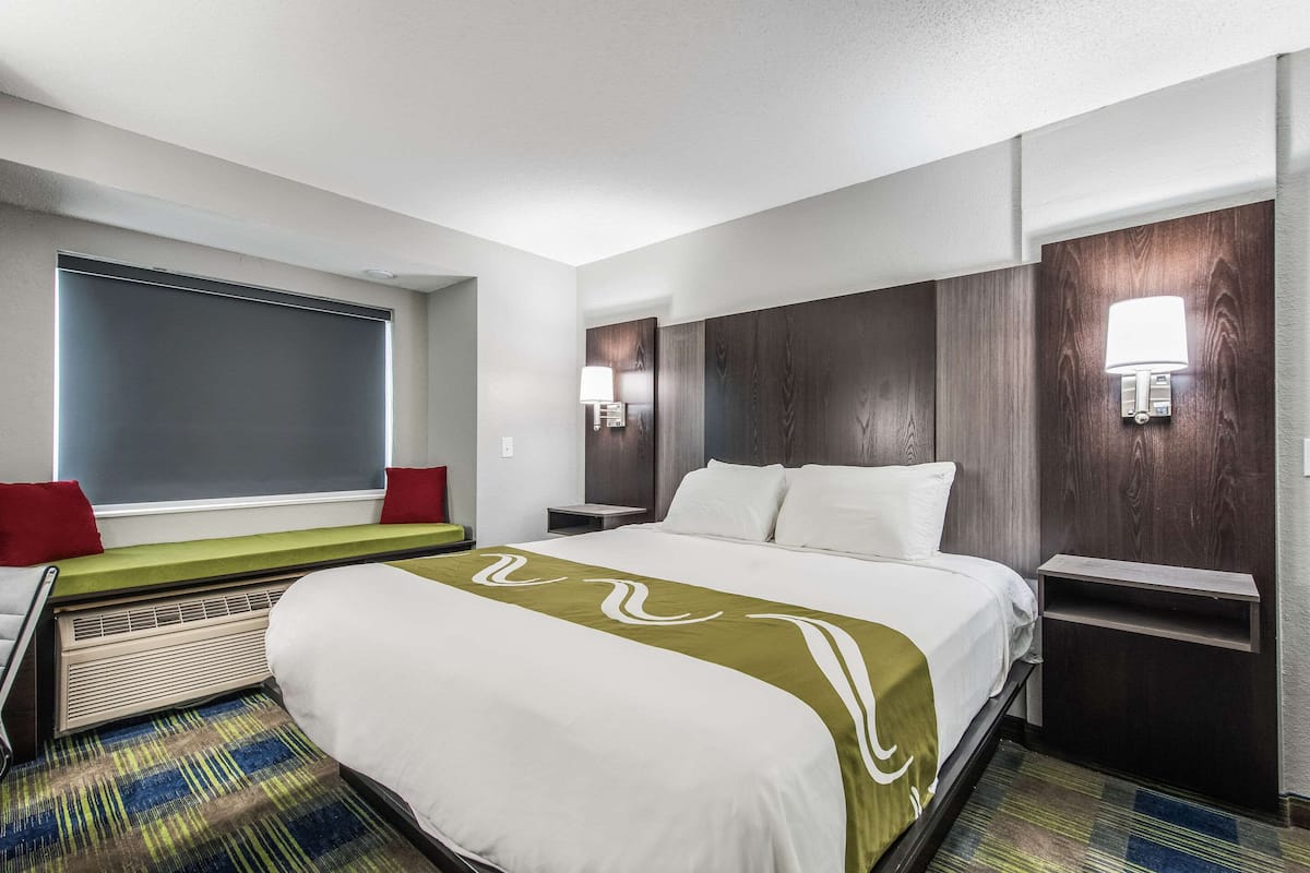 Ein Hotelzimmer mit einem großen Bett, einem Fenster mit Jalousien, einem Nachttisch mit Lampe und einer kleinen Ablagefläche.