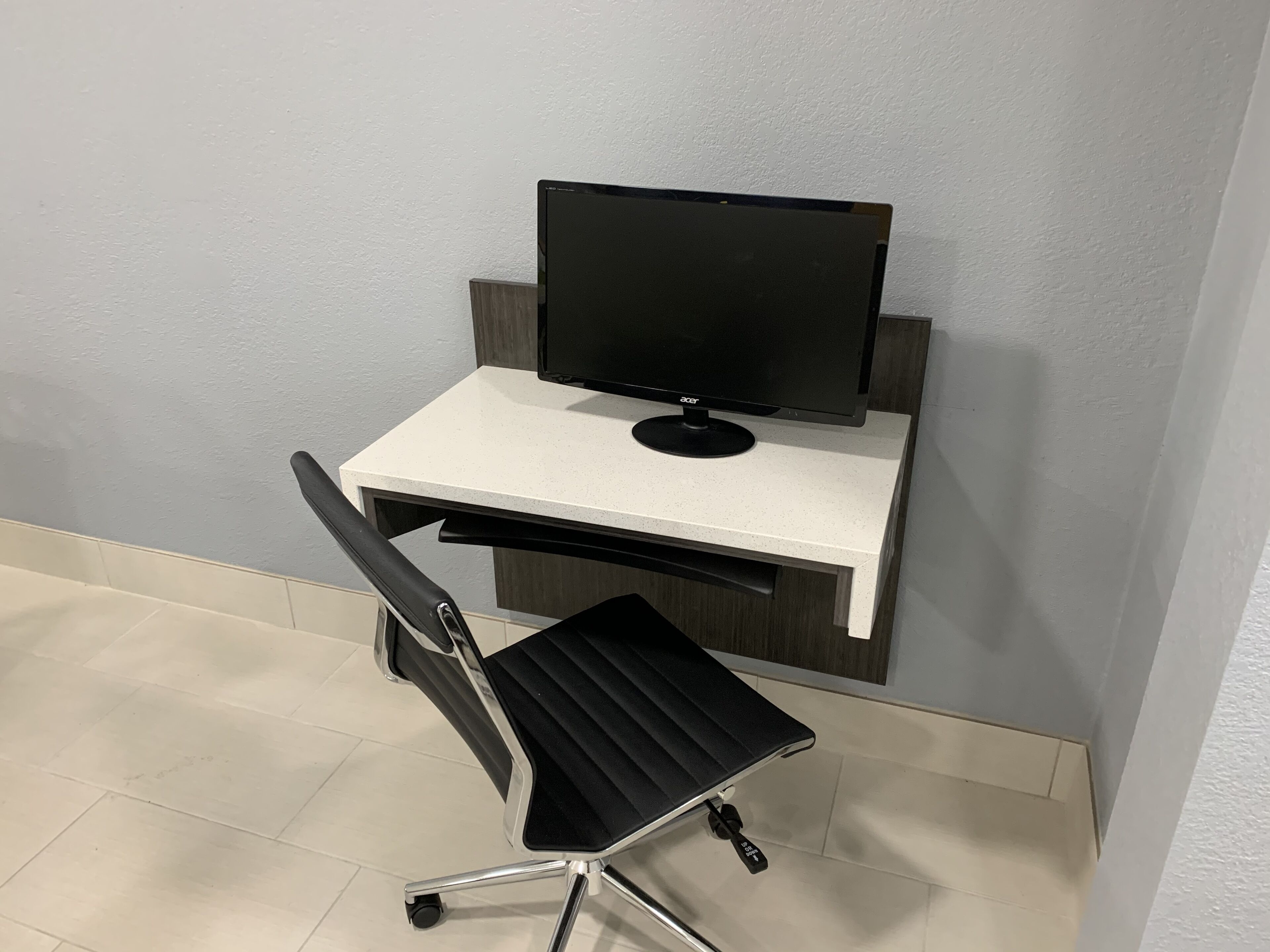 Una moderna scrivania da ufficio con un monitor del computer, una sedia da ufficio nera e un pavimento piastrellato bianco.
