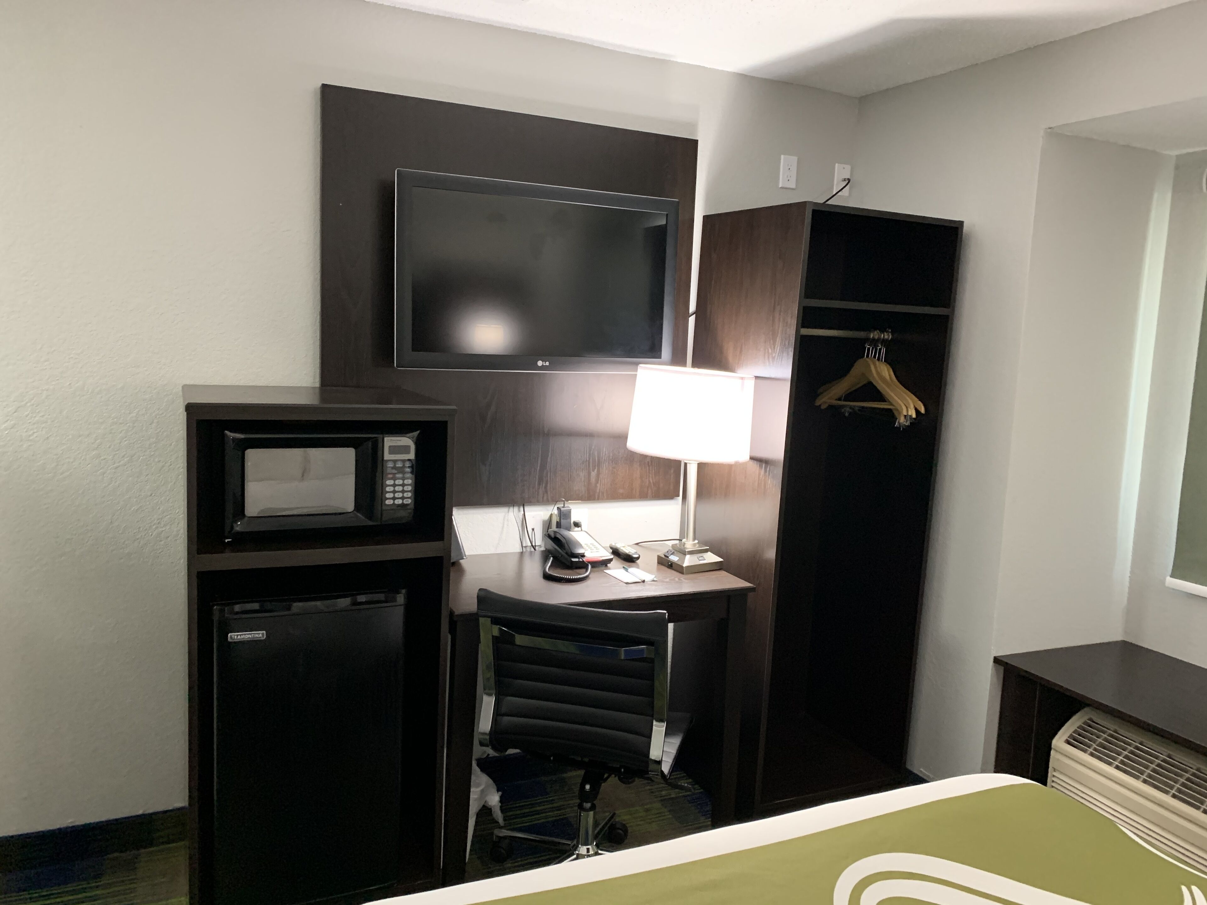 A hotel room with a bed, a desk with a chair, a microwave, a telephone, a TV, and a wardrobe.