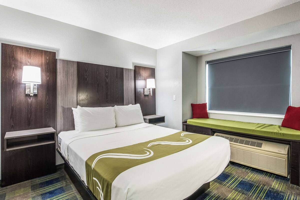 Ein Hotelzimmer mit einem großen Bett, zwei Nachttischen, einer Bank mit einem grünen Kissen, einem Fenster mit Jalousien und Wandleuchten.