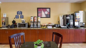 A hotel kitchen with a microwave, coffee machine, and juicer. There are wooden cabinets and a table with a plant.