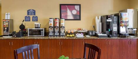 Une cuisine d’hôtel équipée d’un four à micro-ondes, d’une machine à café et d’un extracteur de jus. On y trouve des placards en bois et une table agrémentée d’une plante.