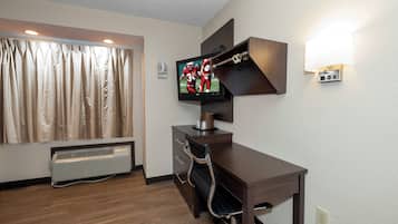 A hotel room with a flat-screen TV mounted on a wall, a desk with drawers, a chair, a window with curtains, and a wall-mounted light fixture.