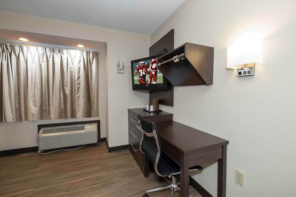 A hotel room with a wooden desk, a flat-screen TV mounted on the wall, a wall-mounted lamp, and a window with curtains.