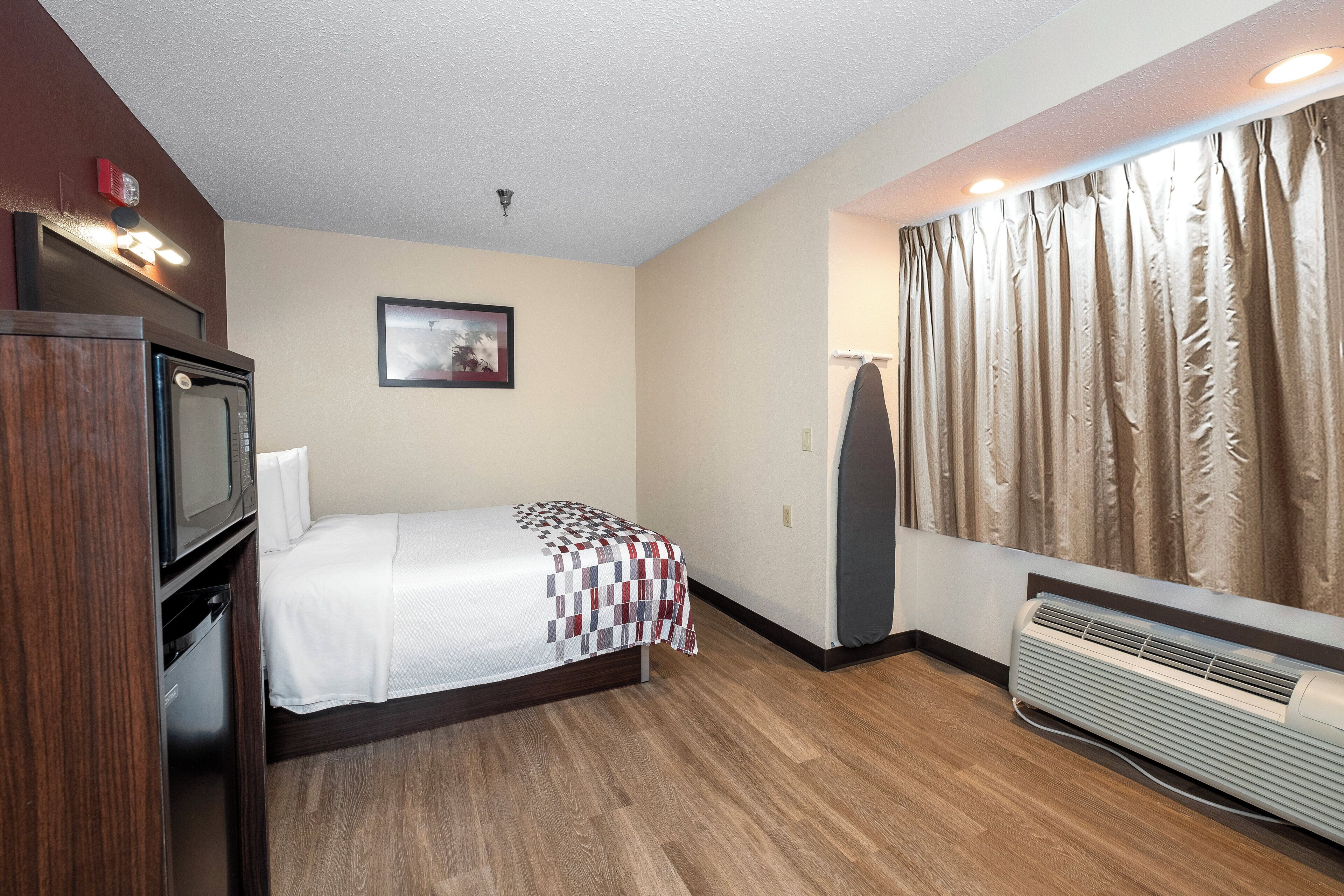 A hotel room with a bed, a television, a picture on the wall, a window with curtains, and a heater.