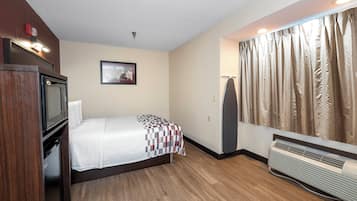 A hotel room with a bed, a television, a picture on the wall, a window with curtains, and a heater.