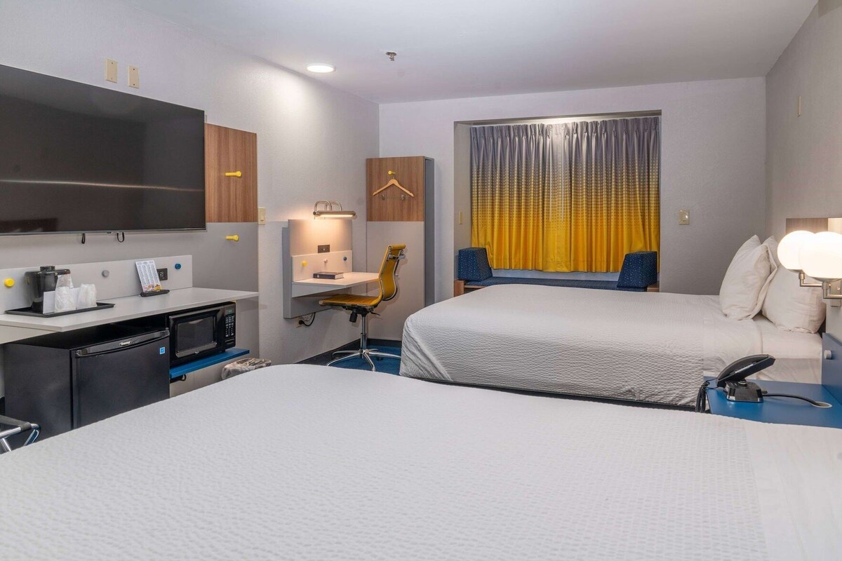 Une chambre d’hôtel avec deux lits, un bureau doté d’un micro-ondes, une chaise, une télévision et une fenêtre avec des rideaux.