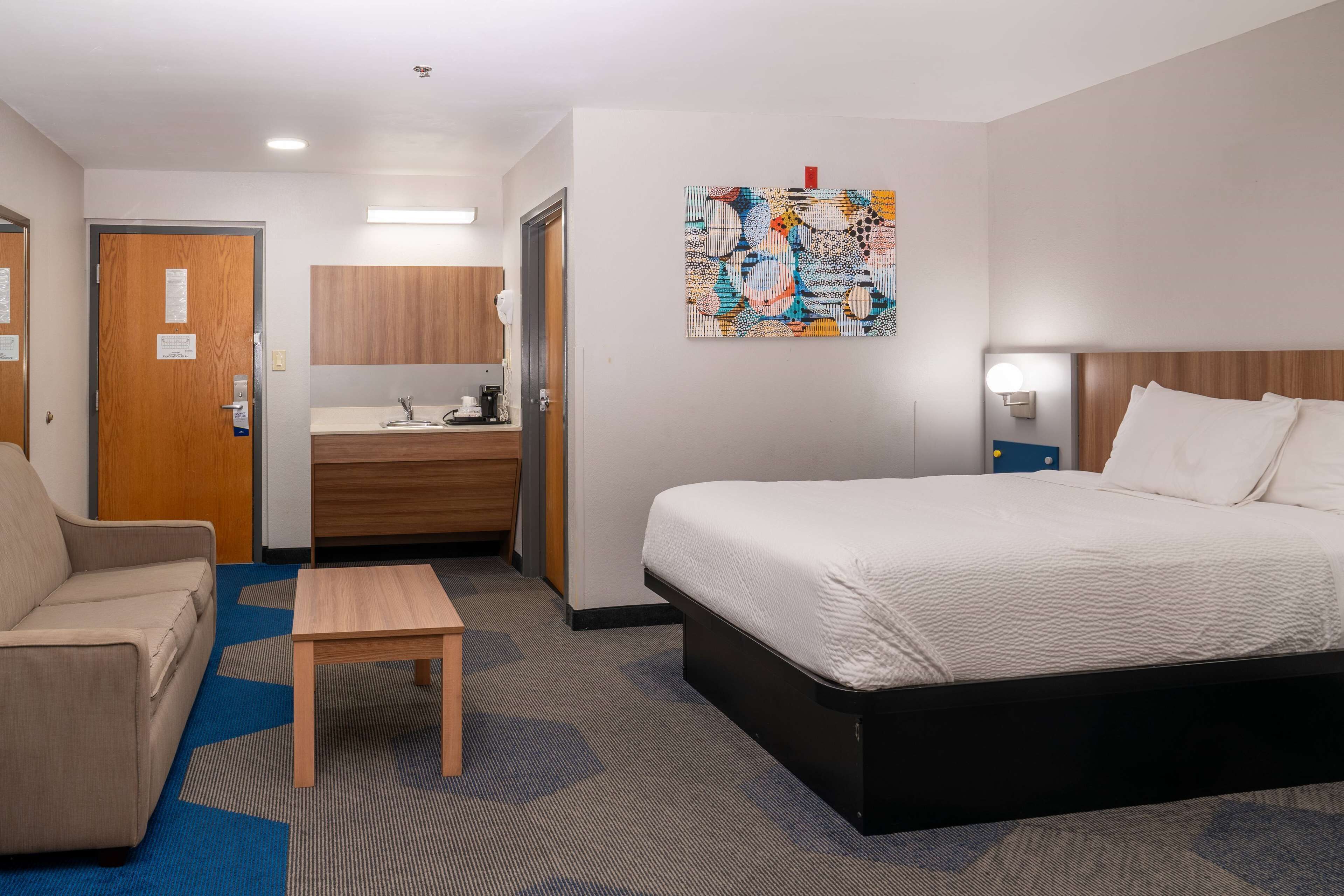 Une chambre d’hôtel comprenant un lit, un canapé, une petite table, une salle de bain avec un lavabo et un miroir, et une peinture abstraite aux couleurs vives accrochée au mur.