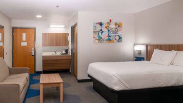 Un cuarto de hotel con una cama, un sofá, una mesita, un baño con lavamanos y espejo, y un cuadro abstracto colorido en la pared.