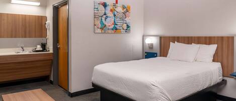 A hotel room with a bed, a wooden nightstand, a small table, a bathroom with a sink and mirror, and a colorful abstract painting on the wall.
